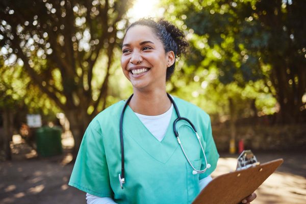 A nurse smiling, holding a clipboard, standing in nature.